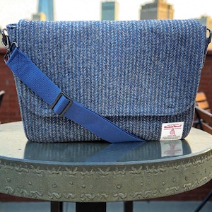 May include: A blue tweed messenger bag with a matching blue shoulder strap. The bag has a flap closure and a small white tag with red text. The bag is sitting on a round, gray table.