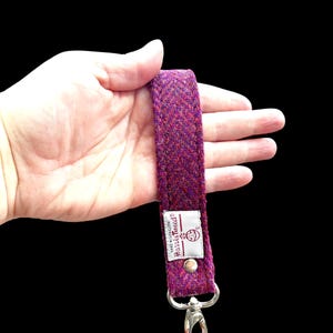 May include: A purple Harris Tweed wrist strap with a silver-colored clasp. The strap is made of woven fabric with a textured appearance. The label reads "Harris Tweed Hand Woven Cloth".