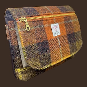 May include: A rectangular clutch bag made of orange, brown, and yellow plaid wool fabric. The bag features a gold-tone metal zipper closure across the front flap and a white rectangular label with the text Harris Tweed printed in black.