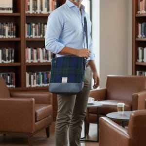May include: A man in a light blue button-down shirt and olive-green pants carries a navy and green plaid messenger bag with a leather bottom. He wears brown loafers. The background includes bookshelves and leather chairs.