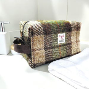 May include: A rectangular toiletry bag made of brown, green, and tan plaid tweed fabric. The bag has a brown handle and a small label. A white towel and a white soap dispenser are in the background.