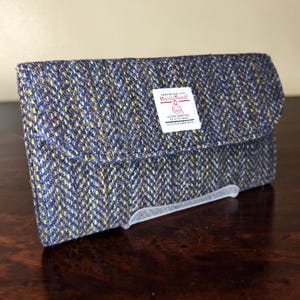 May include: A rectangular wallet made of blue, yellow, and grey herringbone Harris Tweed fabric. A white rectangular label is sewn on the front flap with the text Harris Tweed, Hand Woven Cloth, Outer Hebrides of Scotland, 100% Pure New Wool.