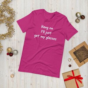 Może przedstawiać: Magenta koszulka z białym napisem "Hang on I'll just get my glasses". Koszulka jest prezentowana na jasnej drewnianej powierzchni z dekoracjami świątecznymi, w tym złotymi i srebrnymi ozdobami oraz zapakowanym prezentem.