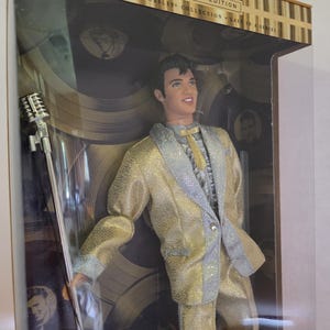May include: A collector edition Elvis Presley doll in a display box. The doll wears a gold and silver textured suit with a ruffled shirt and tie. A silver microphone on a stand is positioned to the left. The box features gold accents and text.