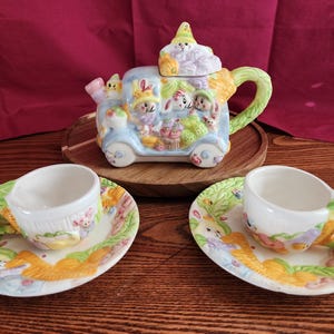 May include: A pastel-colored ceramic tea set featuring a teapot shaped like a bunny-filled wagon. The teapot has a lid with a bunny wearing a hat. The set includes two teacups and saucers with bunny and floral designs.