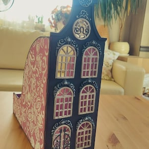 Peut inclure: Un organisateur décoratif en bois en forme de maison. La façade est noire avec des bordures dorées ornées autour des fenêtres et de la porte. Les fenêtres ont un intérieur rose. Le côté de l'organisateur est rose avec un motif floral doré.