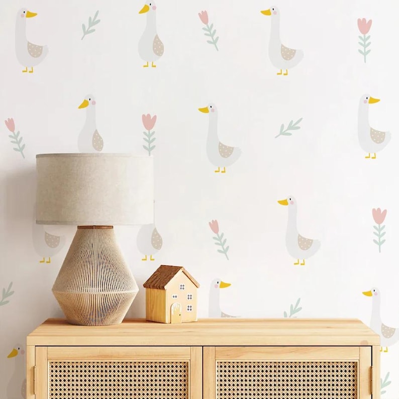 May include: A white wall with a repeating pattern of white geese with yellow beaks and pink flowers. A wooden cabinet with two doors is in the foreground. The cabinet has a light wood finish and a woven rattan front. A lamp with a beige linen shade and a woven base is on the cabinet. A small wooden house is next to the lamp.