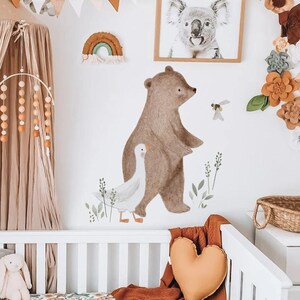 K&ouml;nnte beinhalten: Wandaufkleber f&uuml;r das Kinderzimmer mit einem braunen B&auml;ren und einer wei&szlig;en Gans, mit gr&uuml;nen Bl&auml;ttern. Ein gerahmter Koala-Druck und ein dekorativer Regenbogen sind ebenfalls zu sehen. Die Szene spielt in einem Kinderzimmer mit einem Kinderbett und einem Mobile.