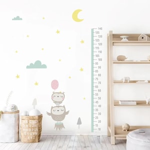 May include: A white wall with a blue and green growth chart featuring a cute owl design. The chart is decorated with yellow stars and clouds. The chart is next to a wooden shelf with a gray ottoman.