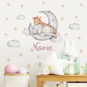 Könnte beinhalten: Ein Wandaufkleber für ein Kinderzimmer mit einem grauen Elefanten und einer Giraffe, die auf einem Halbmond schlafen. Der Aufkleber ist von kleinen weißen Wolken und goldenen Sternen umgeben. Der Text "Name" ist in Rosa unter dem Mond geschrieben.