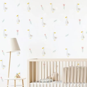 May include: A white crib with a white and gray patterned wallpaper featuring a repeating pattern of white geese and pink flowers. The wallpaper is on a white wall. A white lamp with a white lampshade is on a white side table next to the crib. There is a small wooden toy on the side table.