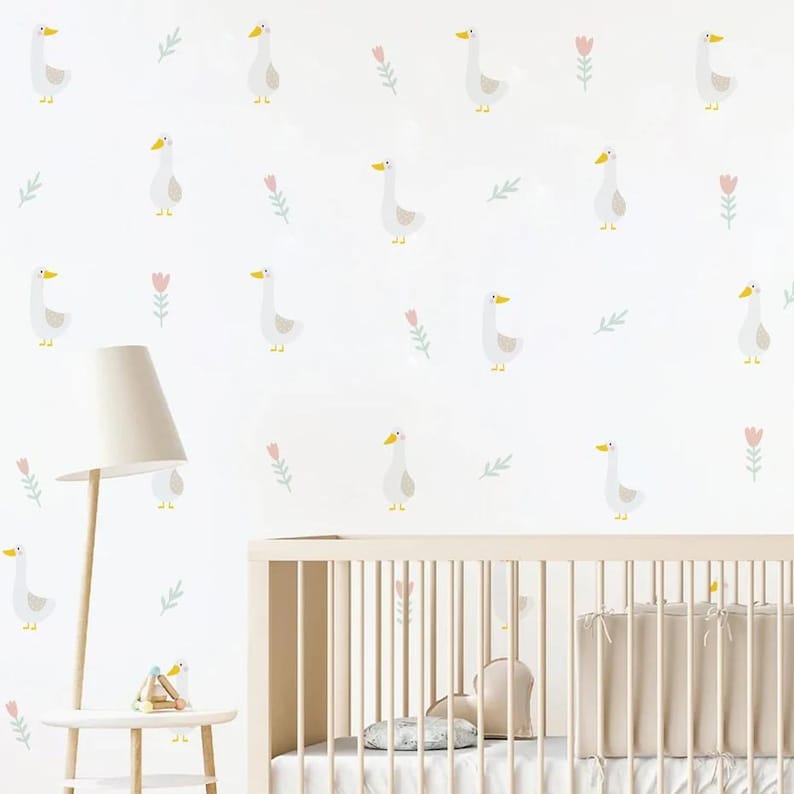 May include: A white crib with a white and gray patterned wallpaper featuring a repeating pattern of white geese and pink flowers. The wallpaper is on a white wall. A white lamp with a white lampshade is on a white side table next to the crib. There is a small wooden toy on the side table.
