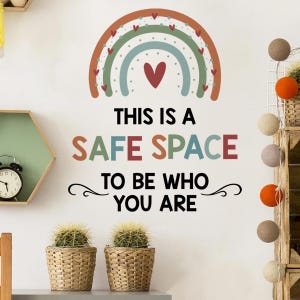 May include: A wall sticker with a rainbow design, a red heart, and the text "THIS IS A SAFE SPACE TO BE WHO YOU ARE." Two small potted cacti and a decorative clock are also visible.