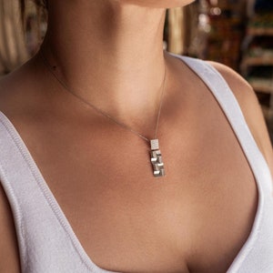 Könnte beinhalten: Eine silberne Halskette mit einem geometrischen Anhänger hängt an einer Person. Die Kette wird über einem weißen Tanktop getragen.