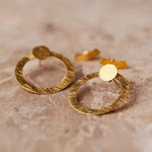 Peut inclure: Une paire de boucles d'oreilles créoles dorées avec une finition texturée. Les boucles d'oreilles ont un design circulaire avec un cercle plus petit au centre.