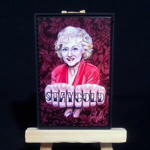 May include: A framed illustration of a woman with blonde hair wearing a red jacket and a red and black background. The woman is holding her hands up with the words "STAY GOLD" tattooed on her knuckles.