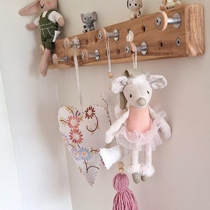May include: A wooden wall-mounted rack with multiple hooks, featuring a variety of stuffed animals, including a bunny, a mouse, a dog, and a kangaroo. The rack is decorated with wooden beads and has a natural wood finish.