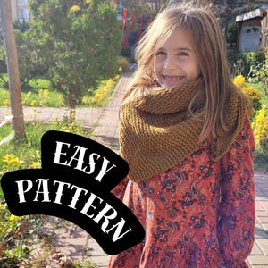 May include: A mustard-colored knitted cowl scarf is worn by a person. The scarf is made of a thick yarn and has a textured appearance. The person is wearing a red dress with a paisley pattern. The words "EASY PATTERN" are in a black bubble.