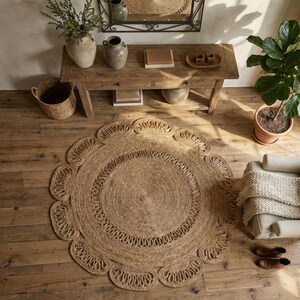 May include: A round, natural fiber rug with a scalloped edge. The rug is made of woven jute and has a circular design in the center. The rug is placed on a wooden floor, with a wooden table, chair, and potted plant in the background.