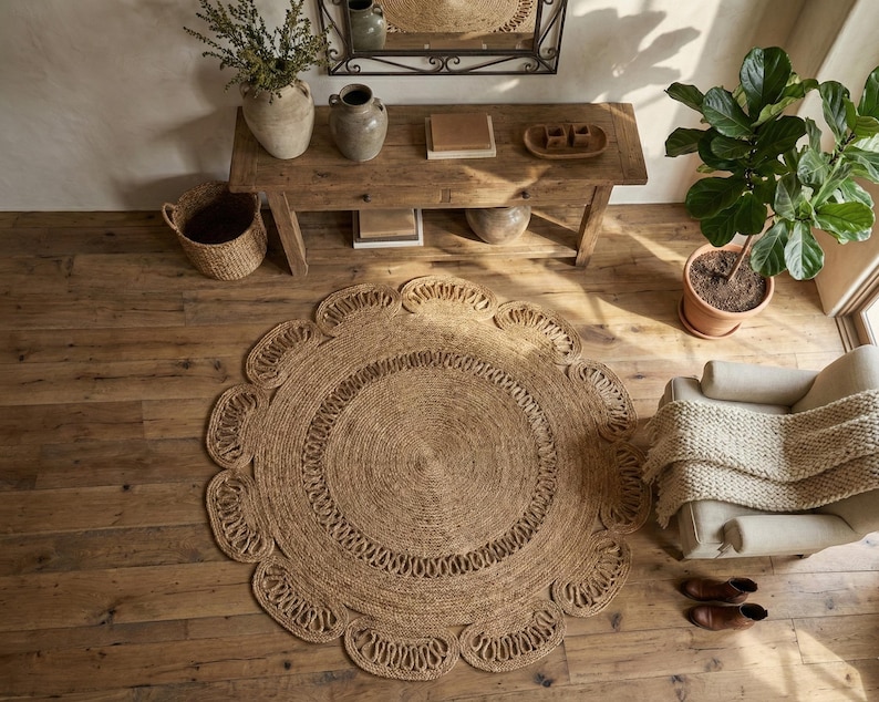 May include: A round, natural fiber rug with a scalloped edge. The rug is made of woven jute and has a circular design in the center. The rug is placed on a wooden floor, with a wooden table, chair, and potted plant in the background.