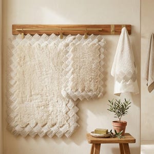 May include: A bathroom set featuring a cream-colored bath mat and a smaller matching rug, both with a textured, scalloped edge. A wooden towel rack with brass accents holds a matching towel. A small wooden stool holds a potted plant and soap.