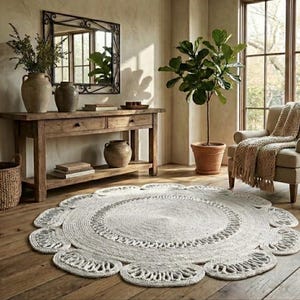 May include: A neutral-toned living room with a large, round, white jute rug with a floral design. The room features a wooden console table, a mirror, a potted plant, and an armchair with a fringed throw blanket. The floor is made of wooden planks.