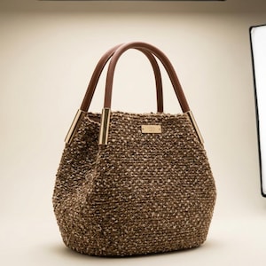 Puede incluir: Un bolso tote marr&oacute;n con un dise&ntilde;o tejido texturizado. El bolso presenta asas de cuero marr&oacute;n y detalles met&aacute;licos dorados. Una peque&ntilde;a placa rectangular dorada est&aacute; centrada en la parte delantera del bolso.