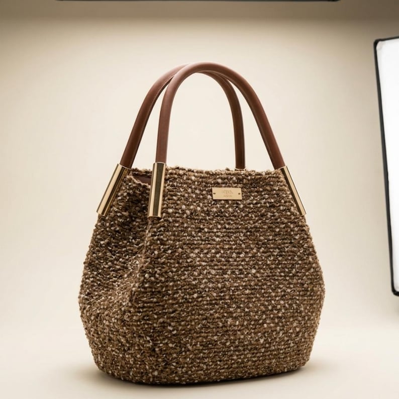 Puede incluir: Un bolso tote marr&oacute;n con un dise&ntilde;o tejido texturizado. El bolso presenta asas de cuero marr&oacute;n y detalles met&aacute;licos dorados. Una peque&ntilde;a placa rectangular dorada est&aacute; centrada en la parte delantera del bolso.