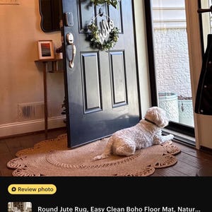 May include: A round jute rug with a scalloped edge lies on a wooden floor. A white dog rests on the rug near a black door adorned with a wreath and a "hello" sign. The rug is a natural beige color.