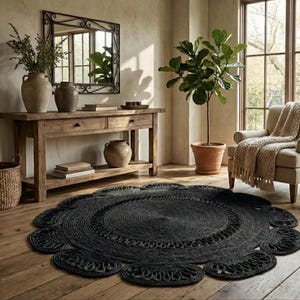 May include: A black, round jute rug with a floral-inspired design. The rug is placed on a wooden floor in a room with a wooden console table, a mirror, and a potted plant. The rug's diameter is approximately 72 inches.