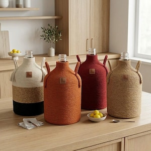 Puede incluir: Cuatro botellas de vidrio decorativas con cubiertas de cuerda tejida en beige, naranja, rojo y marrón claro. Cada botella tiene una etiqueta de cuero y asas. Las botellas se exhiben sobre una superficie de madera, sugiriendo una cocina o comedor.