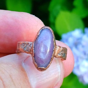 May include: A handcrafted copper ring featuring a large, oval-shaped, lavender-colored gemstone. The ring has a rustic, textured band and bezel setting, showcasing the natural beauty of the stone. The ring is held in a person's hand against a blurred green and blue background.