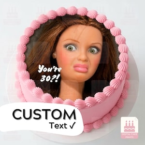 Puede incluir: Un pastel redondo con bordes de glaseado rosa que presenta una imagen comestible central de una cara de muñeca con cabello castaño y ojos verdes. El texto en la imagen dice You're 30?!. El pastel tiene un acabado lateral rosa liso.