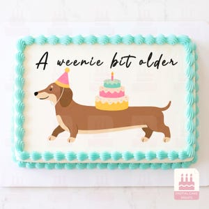 Könnte beinhalten: Ein rechteckiger Blechkuchen mit weißer Glasur und hellblauen Spritzrändern. Oben ist ein brauner Dackel mit rosa Partyhut abgebildet, der eine dreistöckige Torte auf dem Rücken trägt, sowie der schwarze Text A weenie bit older.