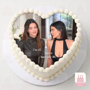 Könnte beinhalten: Ein herzförmiger Kuchen mit weißem Spritzglasur-Rand und einem aufgedruckten Bild von zwei Personen. Der Text auf dem Bild lautet, I'm 27. You're basically 30. Der Kuchen wird auf einer runden weißen Unterlage mit einem kleinen Logo präsentiert.