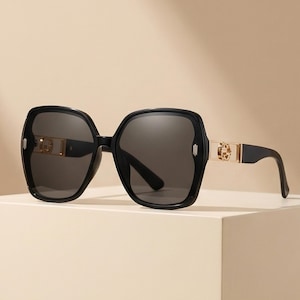 May include: Black oversized sunglasses with dark gray lenses. The frames are a glossy black with gold-colored accents on the temples. The sunglasses have a square shape with rounded edges, offering a stylish and modern look.