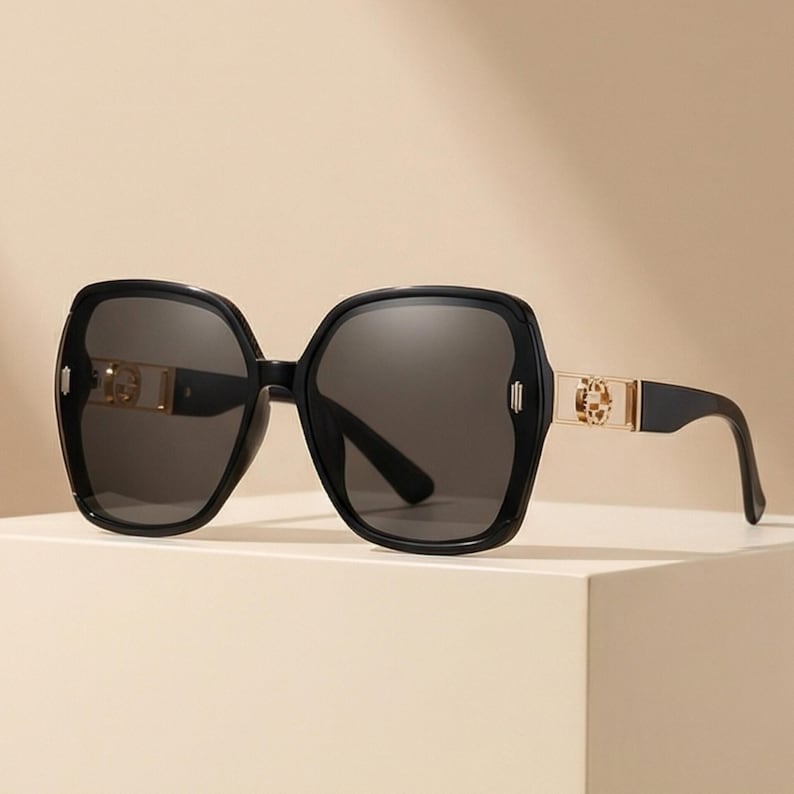 May include: Black oversized sunglasses with dark gray lenses. The frames are a glossy black with gold-colored accents on the temples. The sunglasses have a square shape with rounded edges, offering a stylish and modern look.