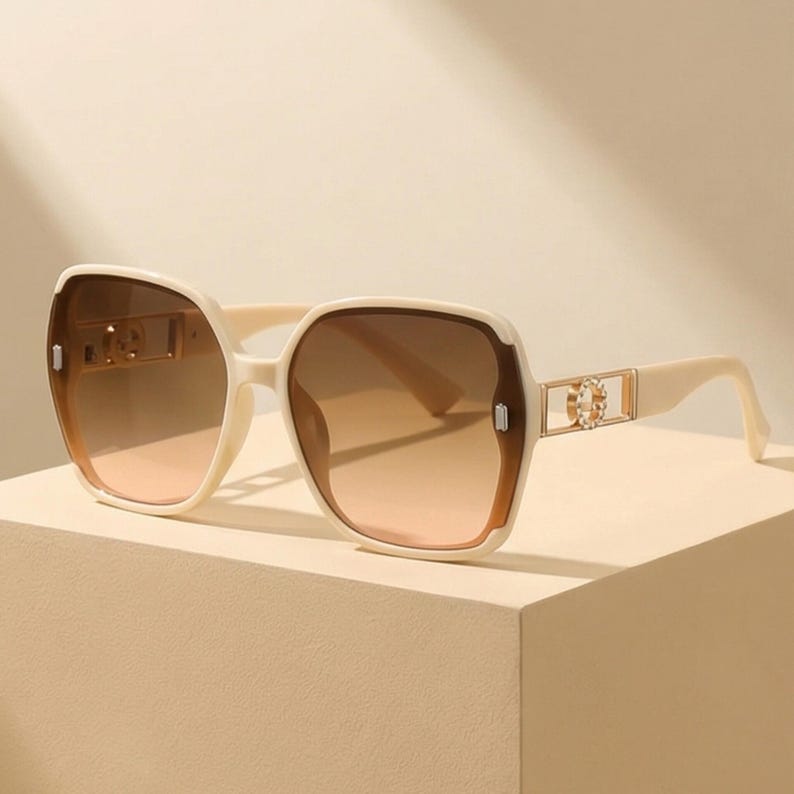May include: Cream-colored sunglasses with gradient brown lenses. The square frames have a thick, rounded design with gold-colored accents on the temples. The lenses transition from a darker brown at the top to a lighter peach color at the bottom.