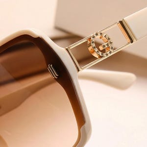 May include: Close-up of oversized sunglasses with a white and brown frame. The sunglasses feature a gold-colored accent with a decorative 'G' logo. The lenses have a gradient tint, transitioning from brown to a lighter shade.