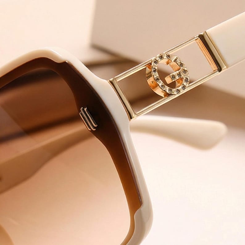 May include: Close-up of oversized sunglasses with a white and brown frame. The sunglasses feature a gold-colored accent with a decorative 'G' logo. The lenses have a gradient tint, transitioning from brown to a lighter shade.