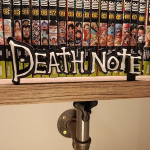 Op de afbeelding: Een zwart-wit "Death Note" bord staat voor een verzameling "One Piece" manga boeken. De boeken staan op een houten plank, ondersteund door een metalen pijp en flens.