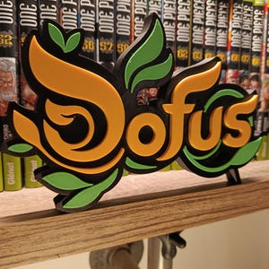 Puede incluir: Logotipo "Dofus" impreso en 3D en naranja, verde y negro, colocado sobre una estantería de madera. El logotipo tiene un diseño estilizado con detalles de hojas. Detrás, varios volúmenes del manga "One Piece".