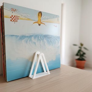 Op de afbeelding: Een stapel vinylplaten met een kleurrijke albumhoes met een persoon die over wolken duikt. Een witte, driehoekige platenhouder ondersteunt de platen op een houten oppervlak.