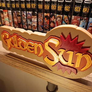 Puede incluir: Un letrero impreso en 3D con las palabras "Golden Sun" en letras doradas y rojas. El letrero está frente a una estantería de libros manga. Los libros son negros con texto blanco e ilustraciones coloridas.