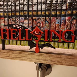 Pode incluir: Um logotipo "Hellsing" vermelho e preto, impresso em 3D, está em frente a uma fileira de volumes de mangá "One Piece". O logotipo apresenta letras estilizadas e um design semelhante a um morcego. As lombadas do mangá são pretas com texto branco e ilustrações coloridas.