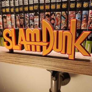 Può includere: Un'insegna "SLAM DUNK" arancione e nera, stampata in 3D, esposta su una mensola di legno. Dietro, una collezione di volumi manga "ONE PIECE". La mensola è sostenuta da una staffa metallica.