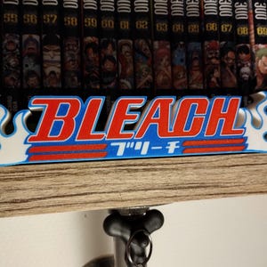 Peut inclure: Une enseigne en 3D imprimée en blanc, bleu et rouge avec le mot "BLEACH" en rouge et le texte japonais "ブリーチ" en noir. L'enseigne est montée sur une étagère en bois avec une rangée de volumes de manga noir et blanc derrière.