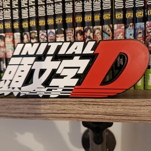 Puede incluir: Logotipo de Initial D impreso en 3D en blanco, negro y rojo, exhibido en una estantería de madera. El logotipo está frente a una colección de volúmenes de manga One Piece. La 'D' roja es prominente.
