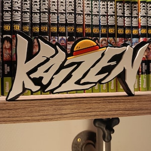 Op de afbeelding: Een wit en zwart "KAZEN" bord met een geel en bruin hoedontwerp, tentoongesteld op een houten plank voor een verzameling "ONE PIECE" manga boeken. Het bord is een decoratief item, waarschijnlijk voor een fan van de mangaserie.