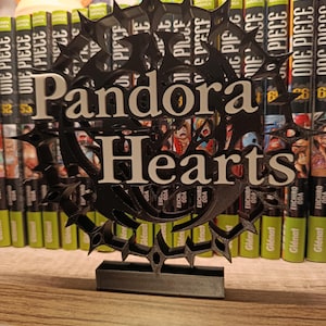 Peut inclure: Un panneau noir et blanc imprimé en 3D avec les mots "Pandora Hearts" dans une police stylisée. Le panneau est placé devant une étagère de mangas, dont des volumes de "One Piece". Le panneau a une bordure décorative et pointue.